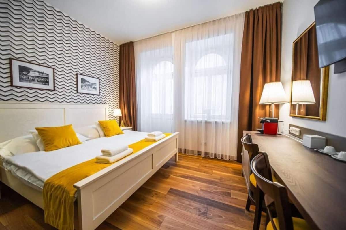 Habitación de hotel con una cama, un escritorio y una silla. Hay dos ventanas con cortinas, una pared con diseño en zigzag y dos lámparas sobre el escritorio.