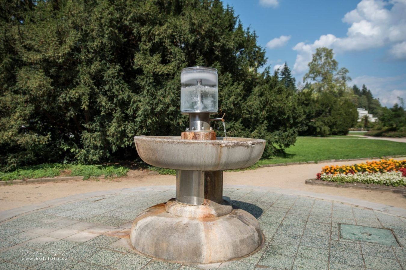 A metal fountain with a water spout, set on a paved area with a grassy background and trees.