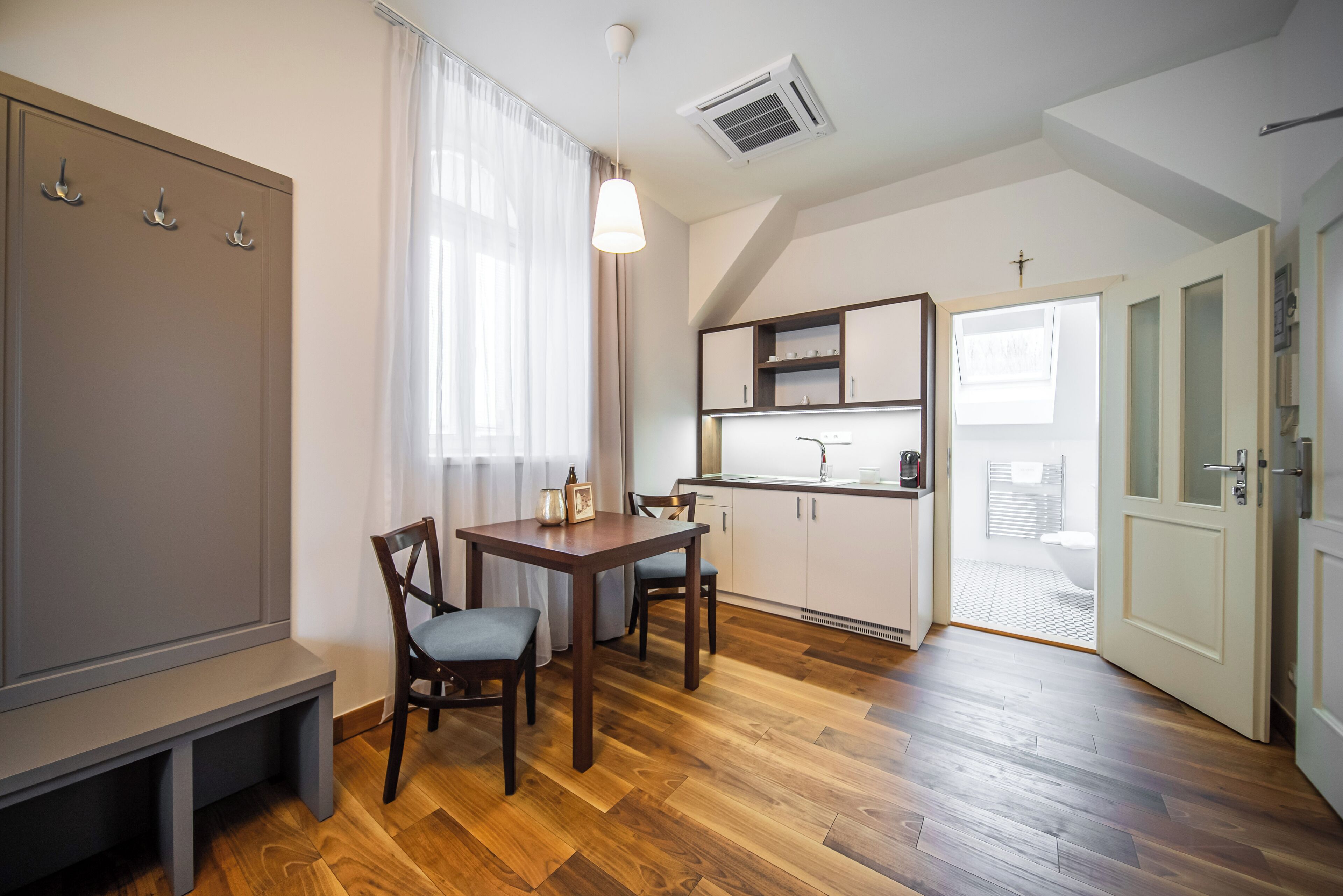 Una habitación compacta con una mesa y sillas de madera, una banca, kitchenette y una puerta que da a un baño.