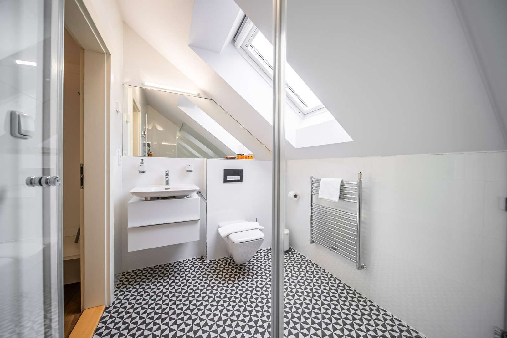 Un baño moderno con piso de diseño geométrico, cabina de ducha de vidrio, lavamanos blanco y un inodoro.