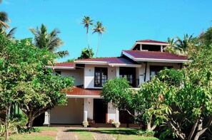 Une maison à deux étages, avec un toit rouge, entourée d’une végétation luxuriante et de palmiers.