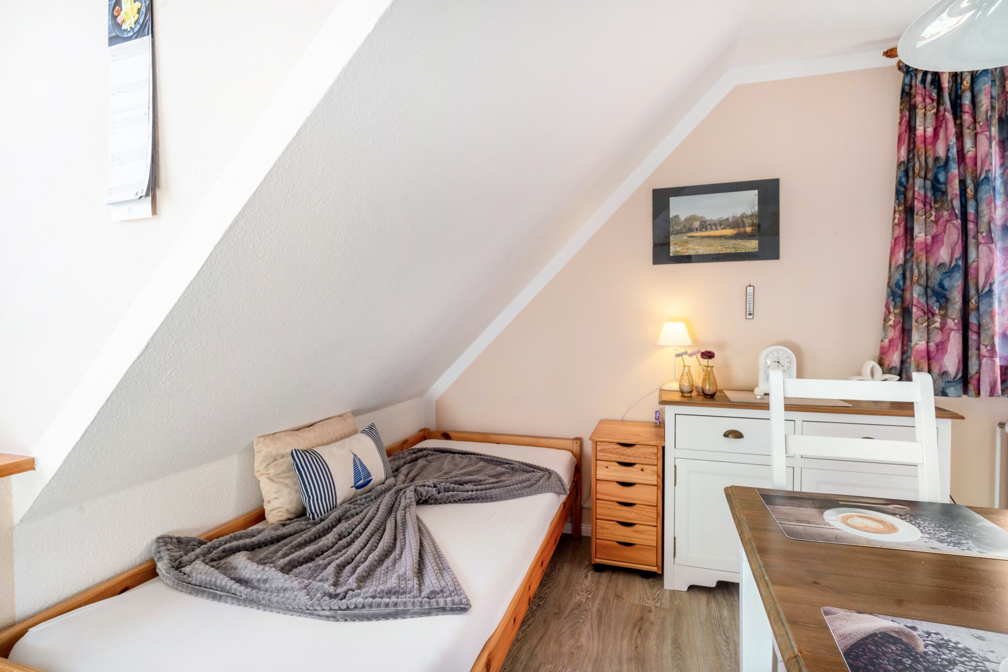 A bedroom with a sloped ceiling, a bed with a blanket, a bedside table with a lamp, a dresser, and a framed picture on the wall.