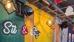 Ein farbenfrohes Interieur mit Neonreklamen mit der Aufschrift "Sit & Sip" und einem Neonherz. Es gibt Pendelleuchten und einen Ventilator an der Decke.
