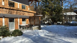 Ein zweigeschossiges Holzhaus mit einem Balkon, einem Essbereich inklusive Stühle und Tisch sowie einem mit Schnee bedeckten Untergrund.