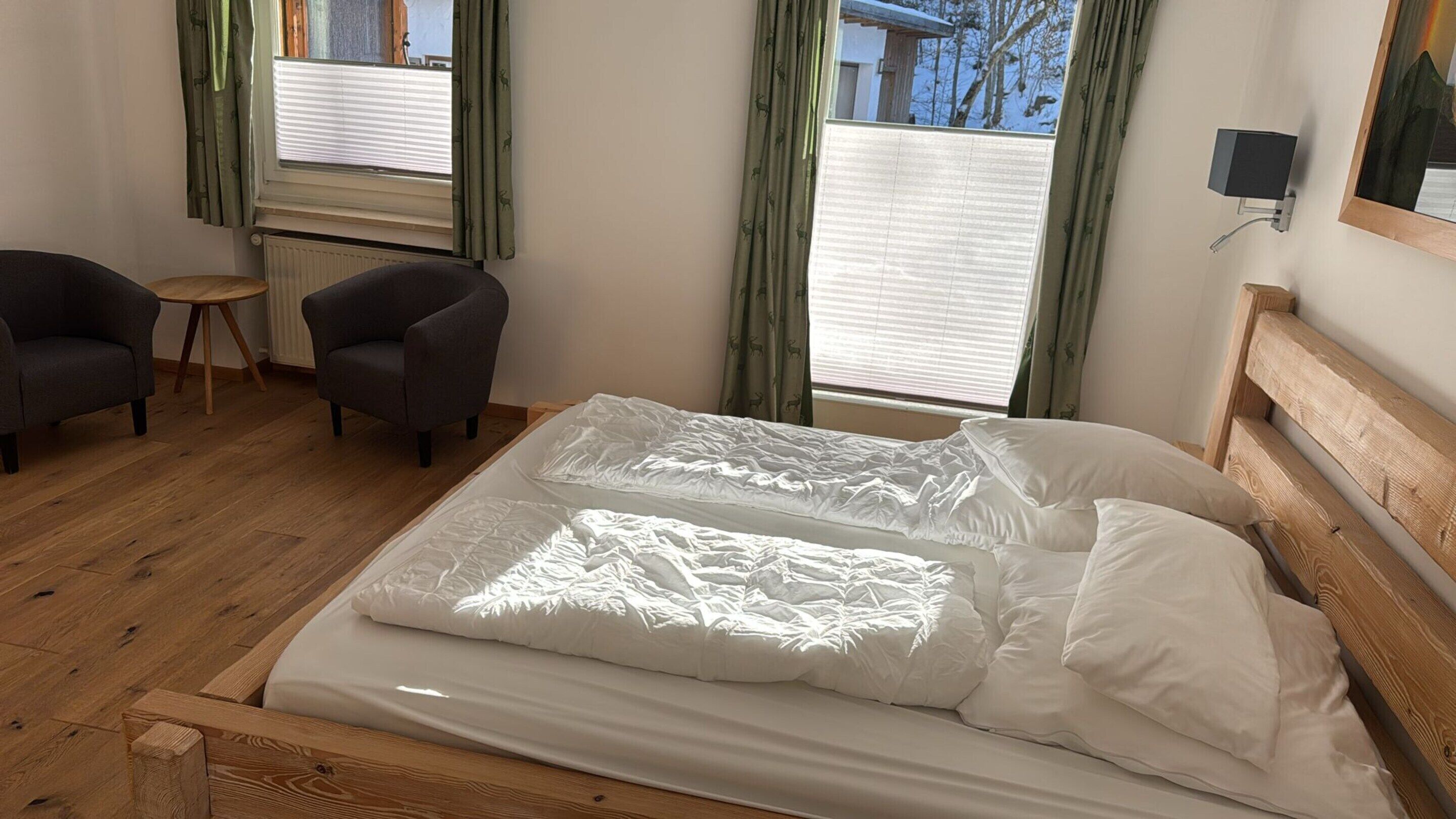 Une chambre à coucher avec un lit en bois, deux fauteuils, une petite table et une fenêtre avec des rideaux.