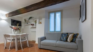 A living room with a grey sofa, white chairs, a tiled floor, a window, and a wooden ceiling beam. - GuestHost - Charming 53sqm apartment, ideal for groups of up to 5 guests, located on the first floor of a historic building without an elevator.Set in the heart of the historic centre, the accommodation is located on one of Ferrara’s most charming medi (Ferrara)