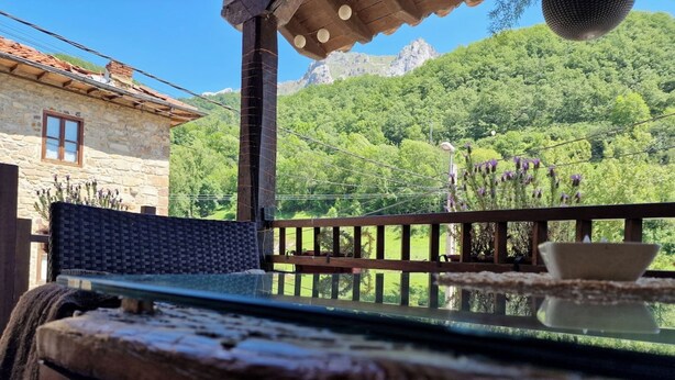 Ein Holztisch mit Glasplatte, ein Korbstuhl und im Hintergrund ein Steinhaus sowie grüne Hügel.