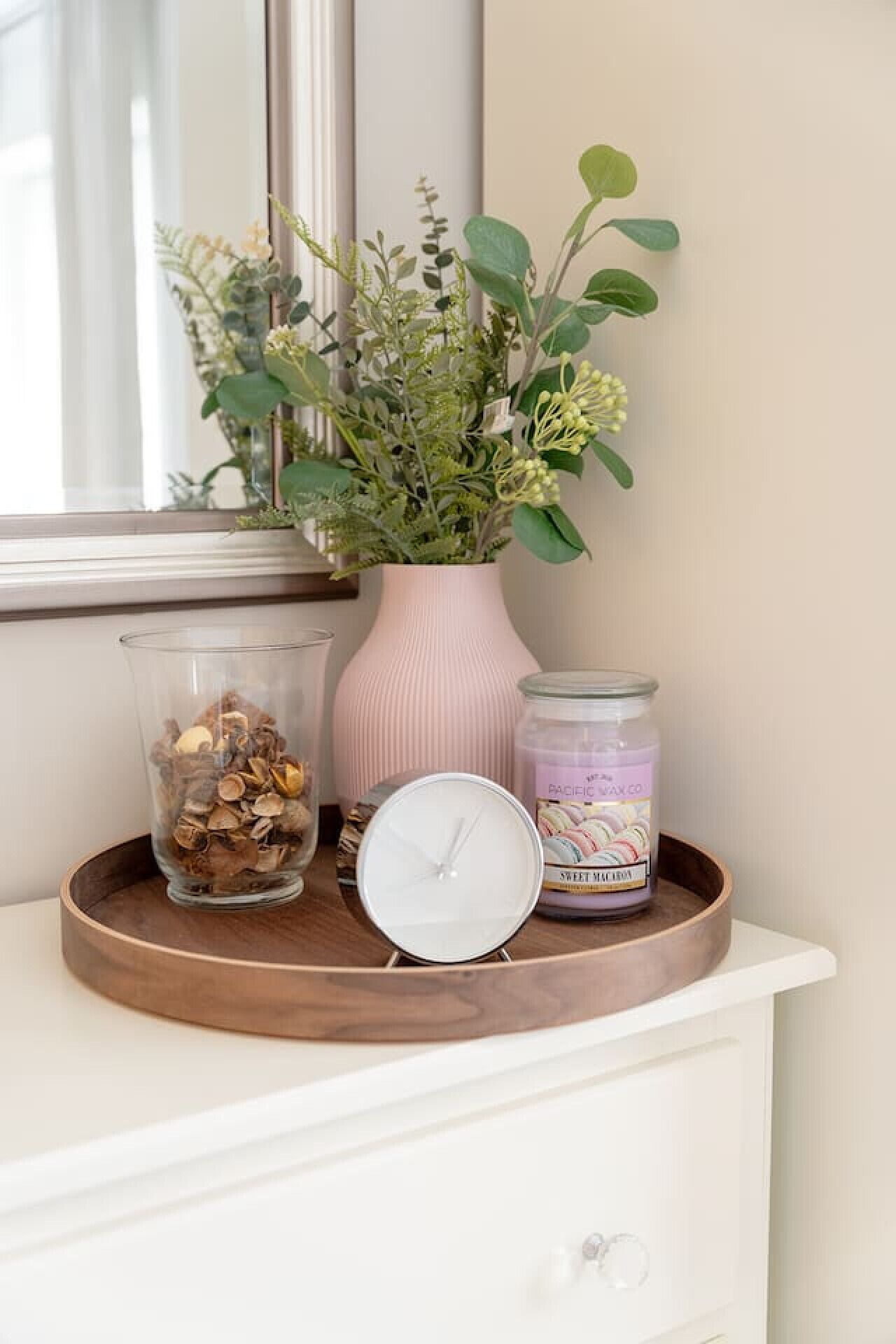 A wooden tray on a white dresser with a clock, a jar of wax paper, and a vase with green plants.
