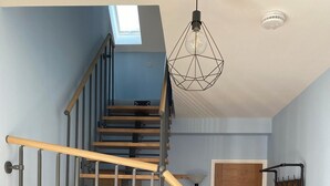 Escada com degraus de madeira e corrimãos de metal, claraboia acima e luminária pendente.