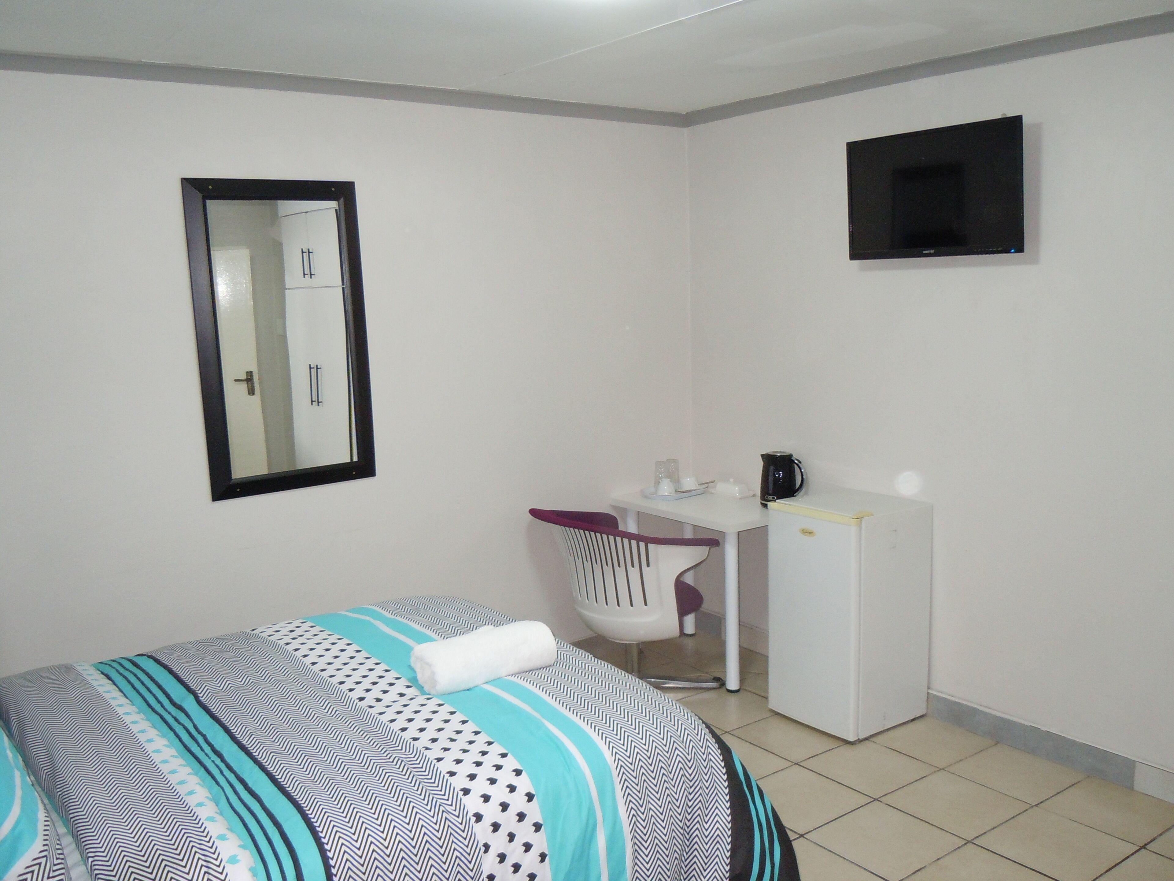 A small, neatly arranged room with a bed, a desk, a chair, a television, and a mirror.