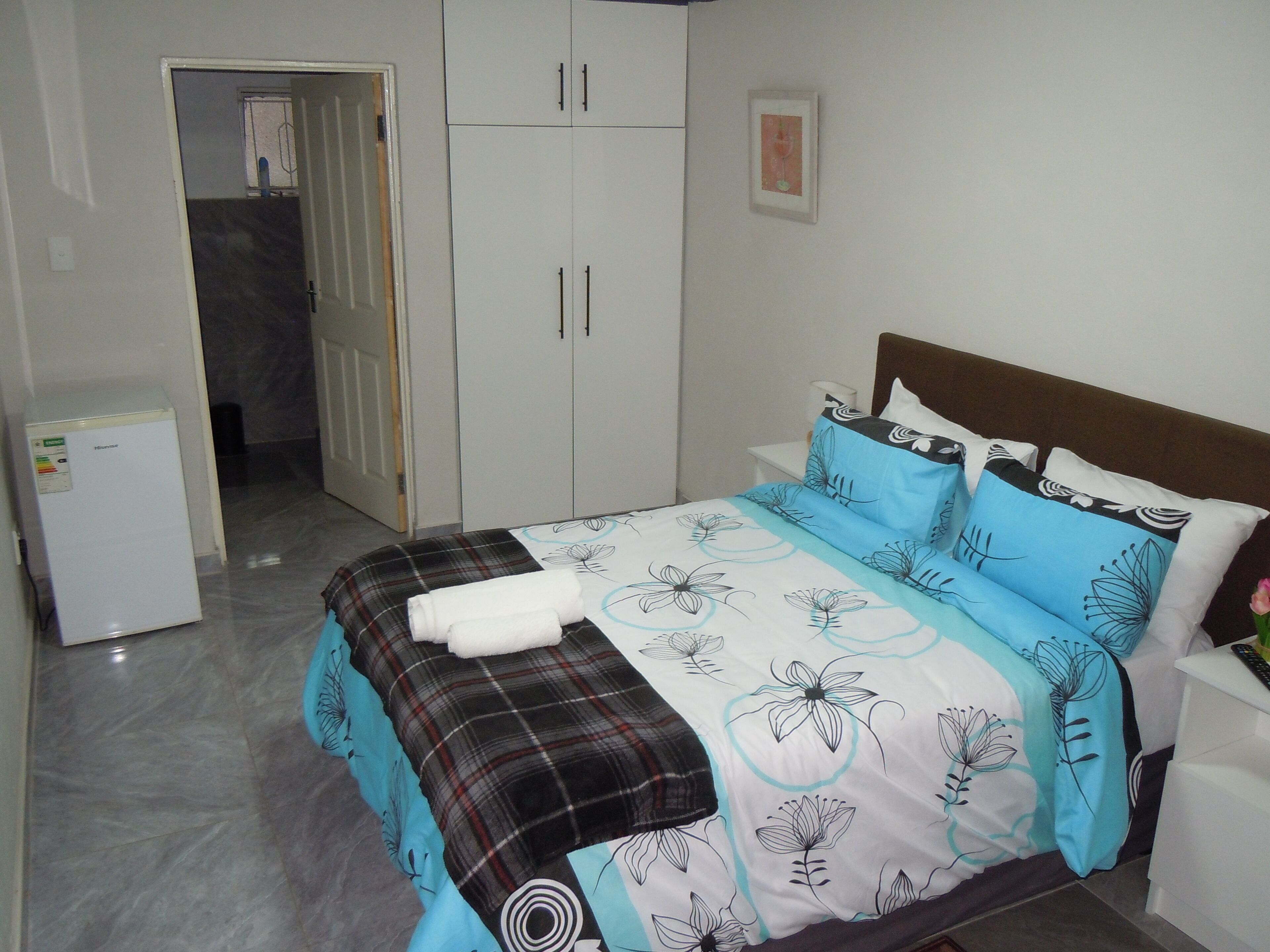 A bedroom with a bed, a wardrobe, a small refrigerator, and a door leading to another room.