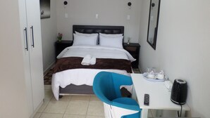A hotel room with a bed, bedside table, chair, and a small table with a remote control and a speaker. - Daeka Guesthouse (Kempton Park)