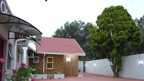 A building with a red-tiled roof, a brick facade, and a sign indicating a reception area. There is a paved area in front with a tree to the right. - Daeka Guesthouse (Kempton Park)