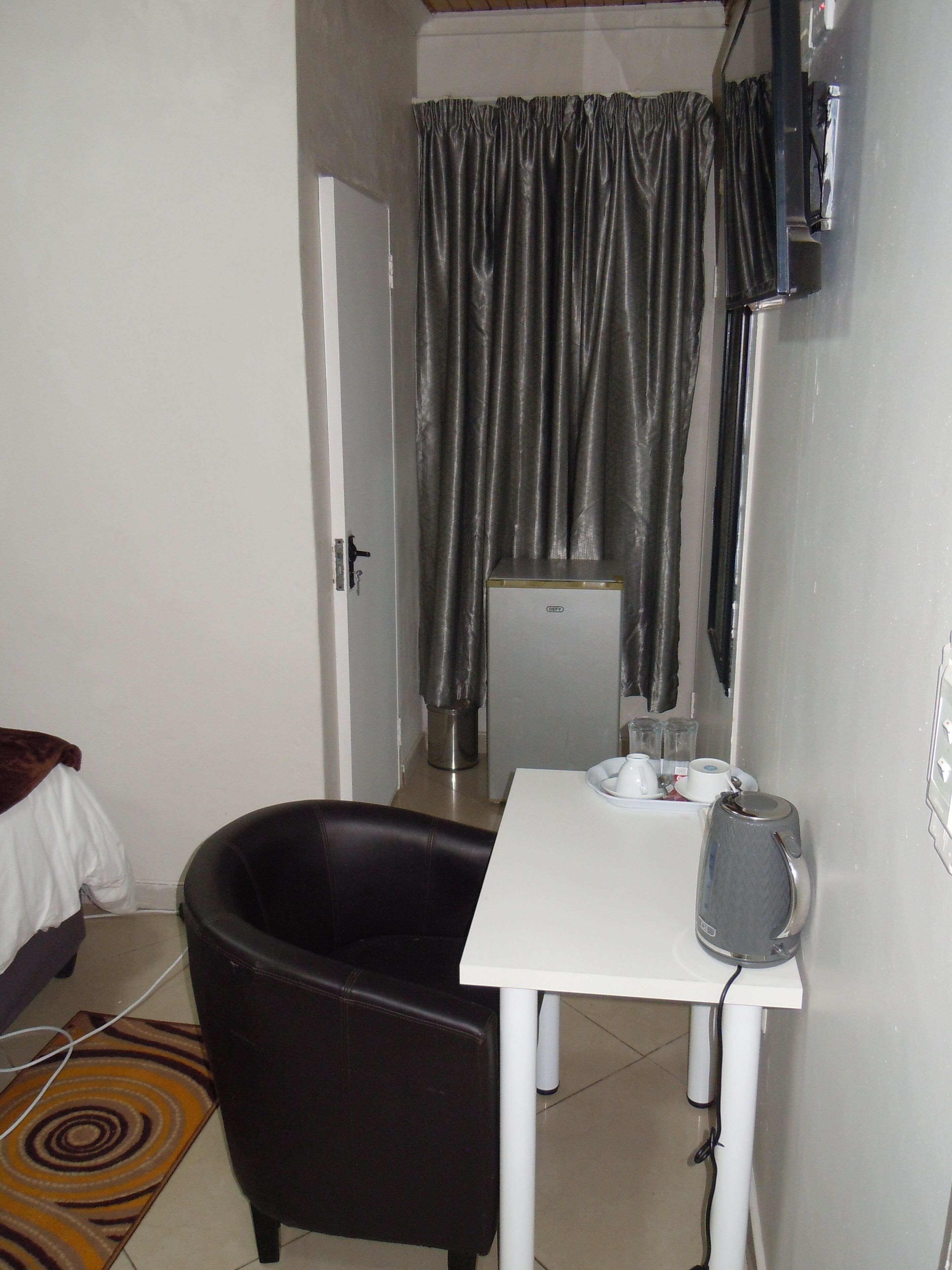 A small room with a black leather chair, a white table with a kettle and cups, a refrigerator, and a curtain.