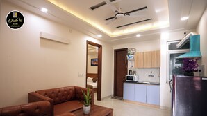 A modern living room with a brown leather sofa, a wooden coffee table, and a potted plant. There is a kitchen area with a microwave, sink, and cabinets. A ceiling fan is visible, and the room has a clean, minimalist design. - Suite 16 Studio Apartment (Gurugram)