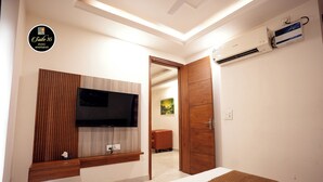 A modern bedroom with a TV mounted on the wall, a bed with a brown rug, and an air conditioning unit on the wall. - Suite 16 Studio Apartment (Gurugram)