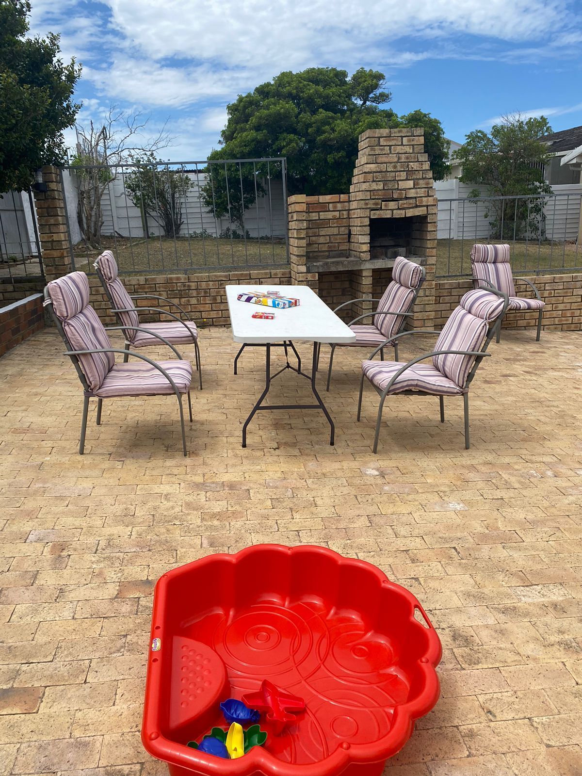 Un patio al aire libre con una mesa de ladrillo y cuatro sillas a rayas, una piscina infantil roja y un horno de ladrillo al fondo.