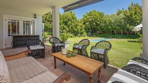 Un patio con mobiliario de mimbre, una mesa de madera y vistas a una piscina y jardín.