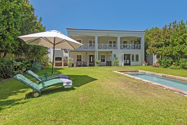 Uma casa de dois andares com piscina, gramado com duas cadeiras de sol e um guarda-sol, e um jardim bem cuidado.