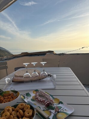 Une table dressée avec des amuse-gueules, une saucisse et des verres à vin, sur une terrasse en plein air offrant une vue sur le paysage.