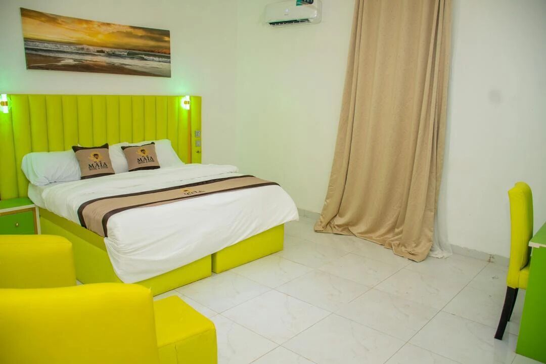 A hotel room with a bed, a green headboard, a yellow chair, and a beige curtain.