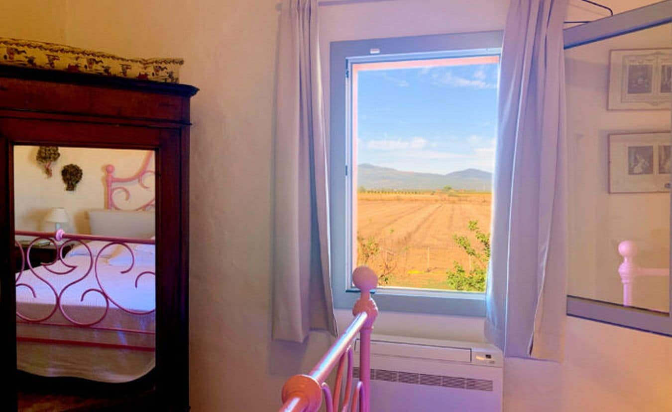 A room with a window overlooking a field, a bed with a decorative headboard, and a small table with a lamp.