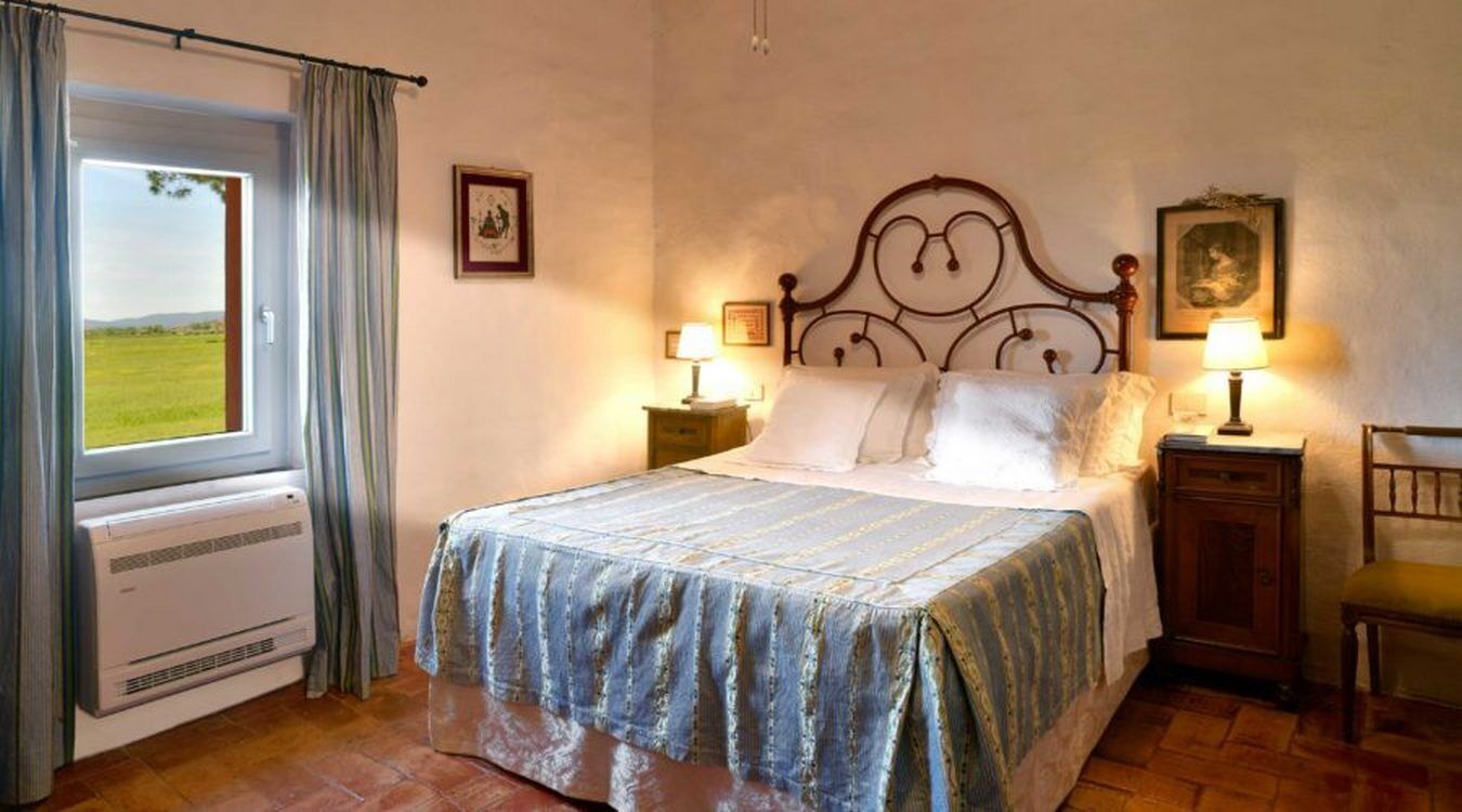 Un dormitorio con cama, dos mesitas de noche con lámparas, una silla, una ventana con cortinas y un radiador.
