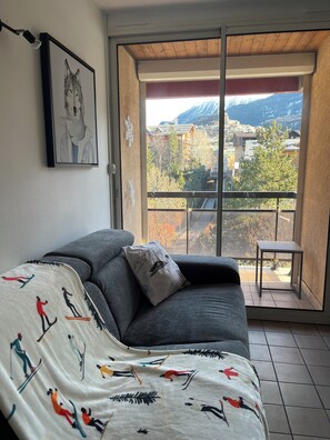 A room with a grey sofa, a blanket with skiing patterns, a framed picture of a wolf, and a view of a mountainous landscape through a large window. - L'Après-Ski, Briançon, at the foot of the Prorel aerial cableway (Briançon)