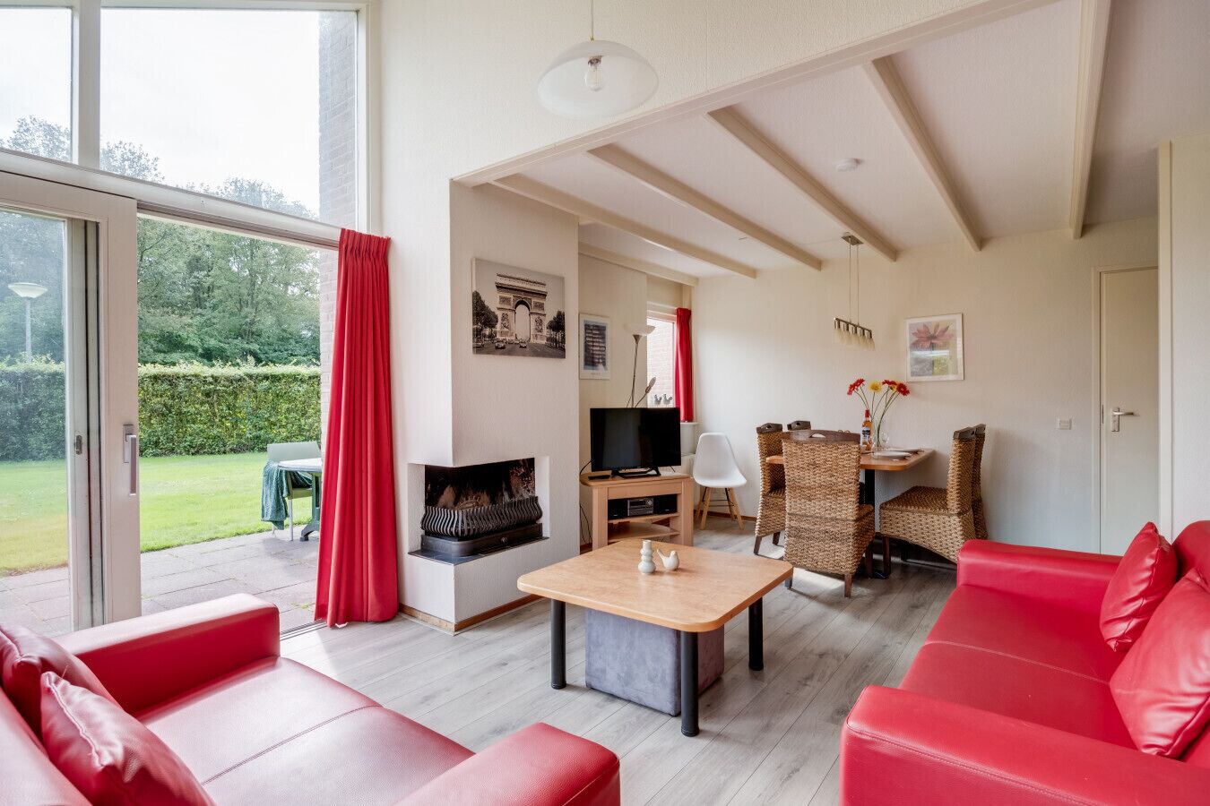 A spacious living room with a red sofa, a wooden coffee table, and a television. There is a dining area with wicker chairs and a table. The room has large windows and a fireplace.
