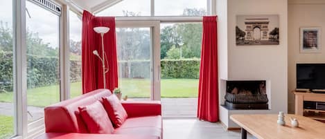 Un salon moderne avec un canapé rouge, une cheminée, un téléviseur et de grandes fenêtres donnant sur l’extérieur.
