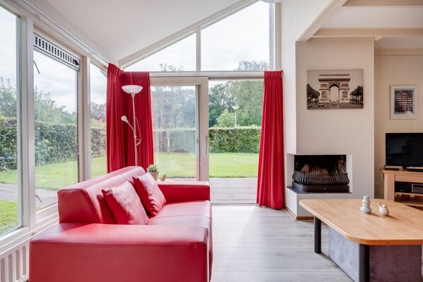 Ein modernes Wohnzimmer mit einem roten Sofa, einem Kamin, einem Fernseher und großen Fenstern mit Blick ins Freie.