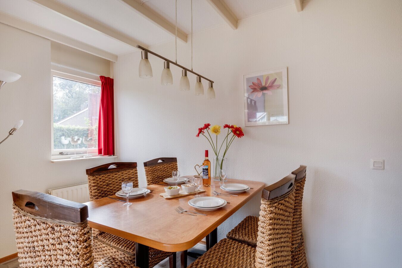 A dining area with a wooden table, woven chairs, and a window with red curtains.