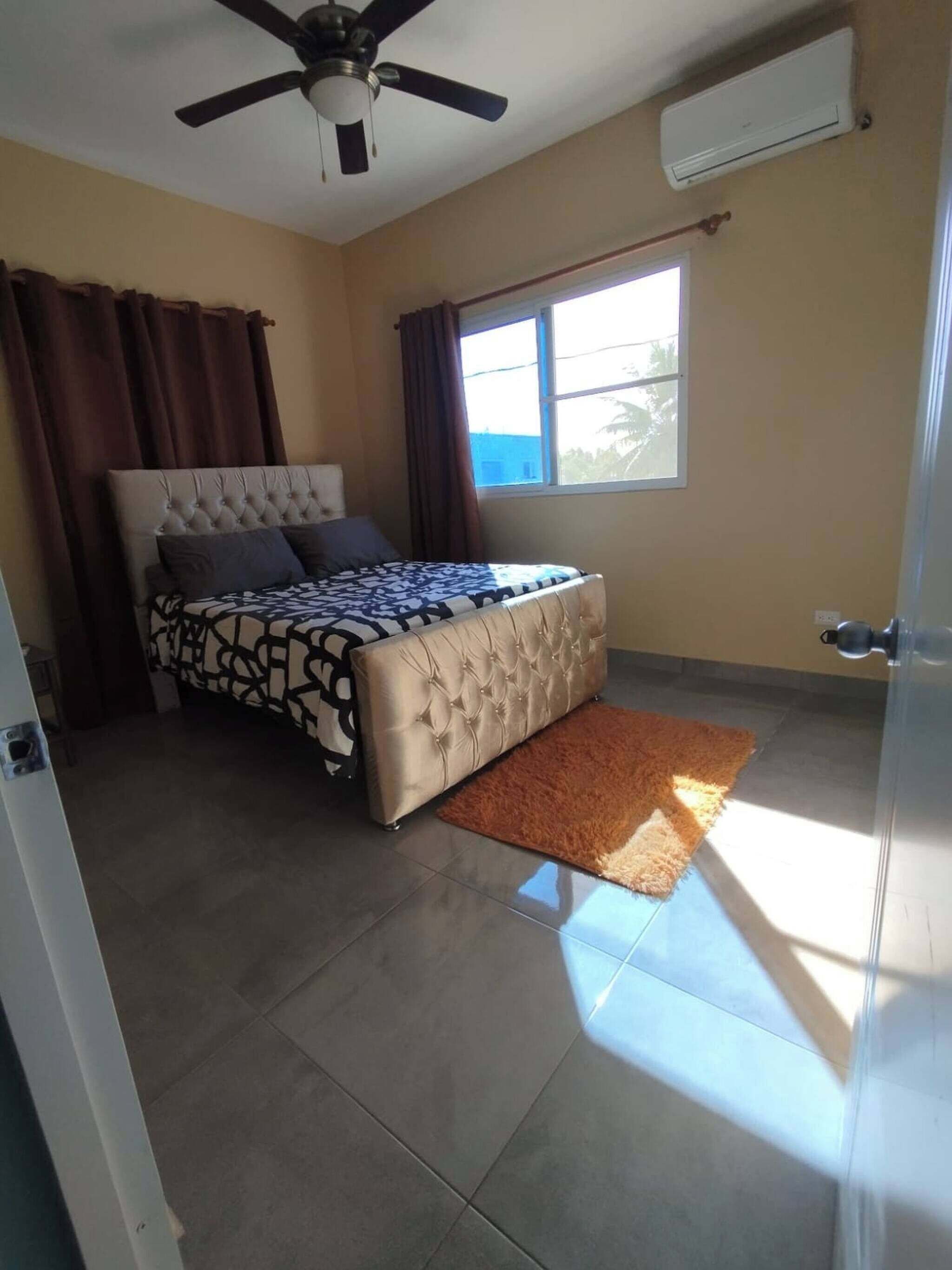 A bedroom with a bed, a ceiling fan, an air conditioning unit, a window, and a door.