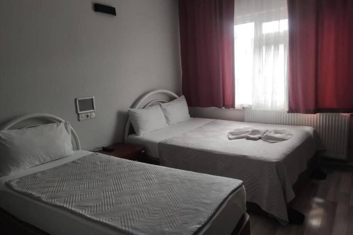 A hotel room with two beds, a bedside table, a nightstand, a window with red curtains, and a wall-mounted thermostat.