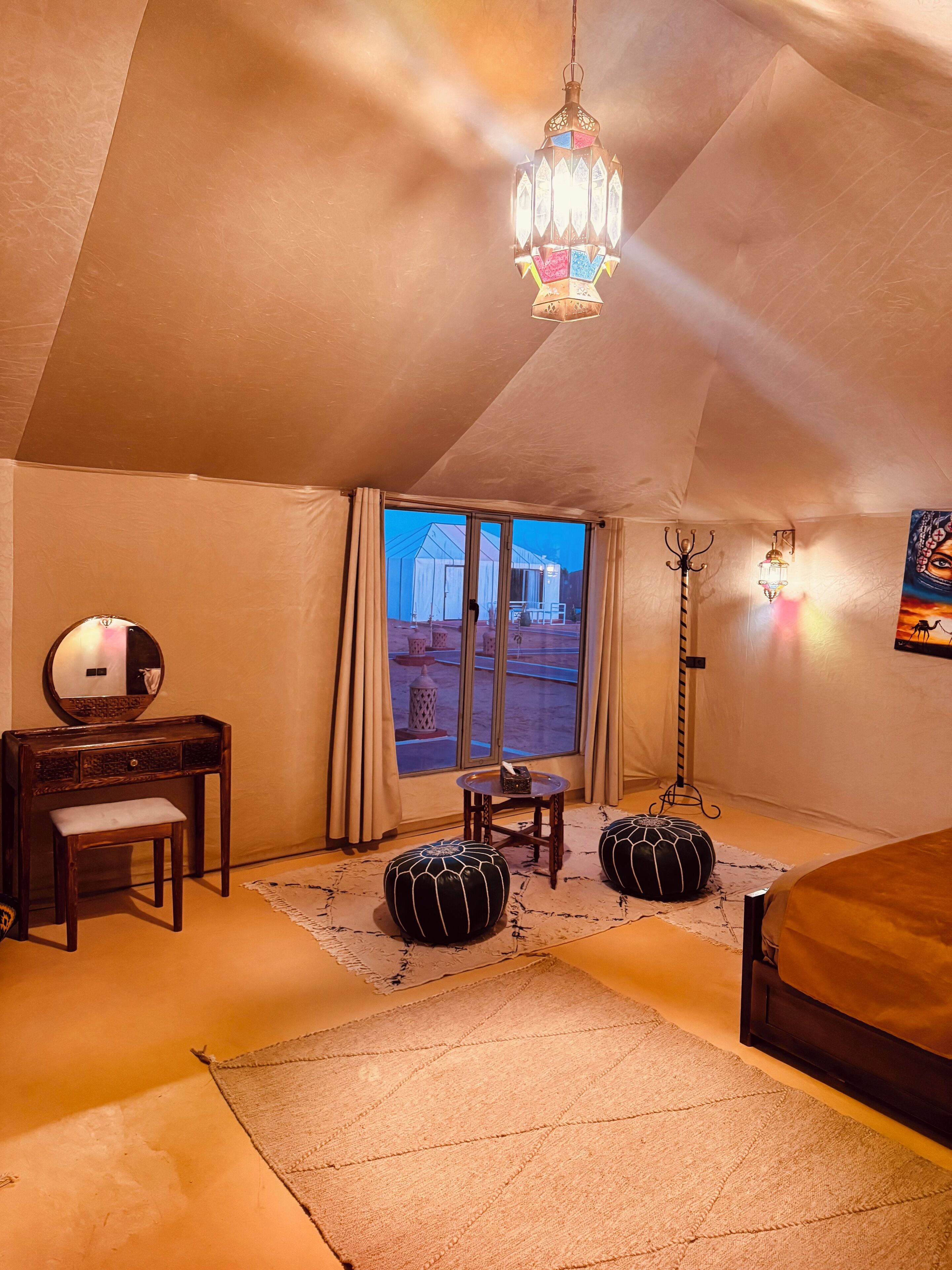 A cozy room with a bed, a vanity with a mirror, a small table, ottomans, and a view of a tented area outside.