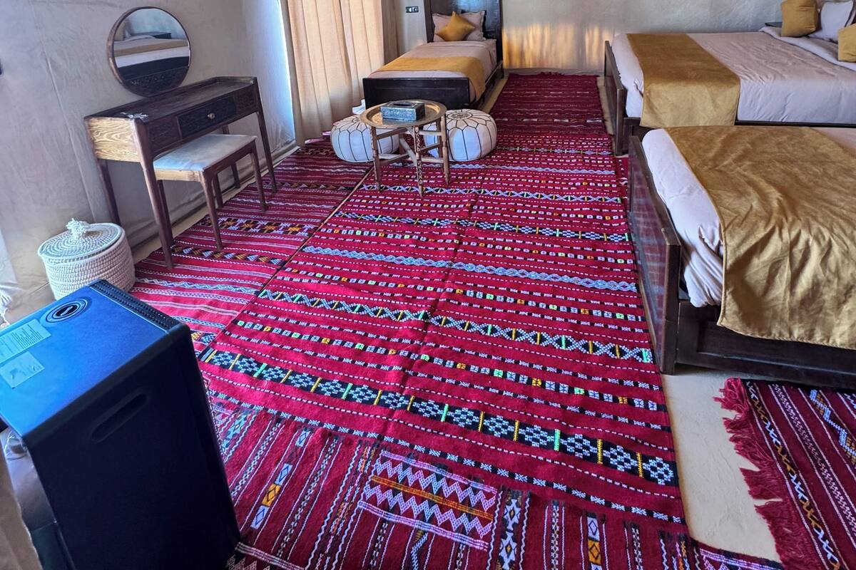 Une chambre mansardée avec un plafond en pente, un lit recouvert d’une couverture dorée, une table de chevet en bois, une petite table avec une lampe, un miroir et un tapis rouge à motifs.