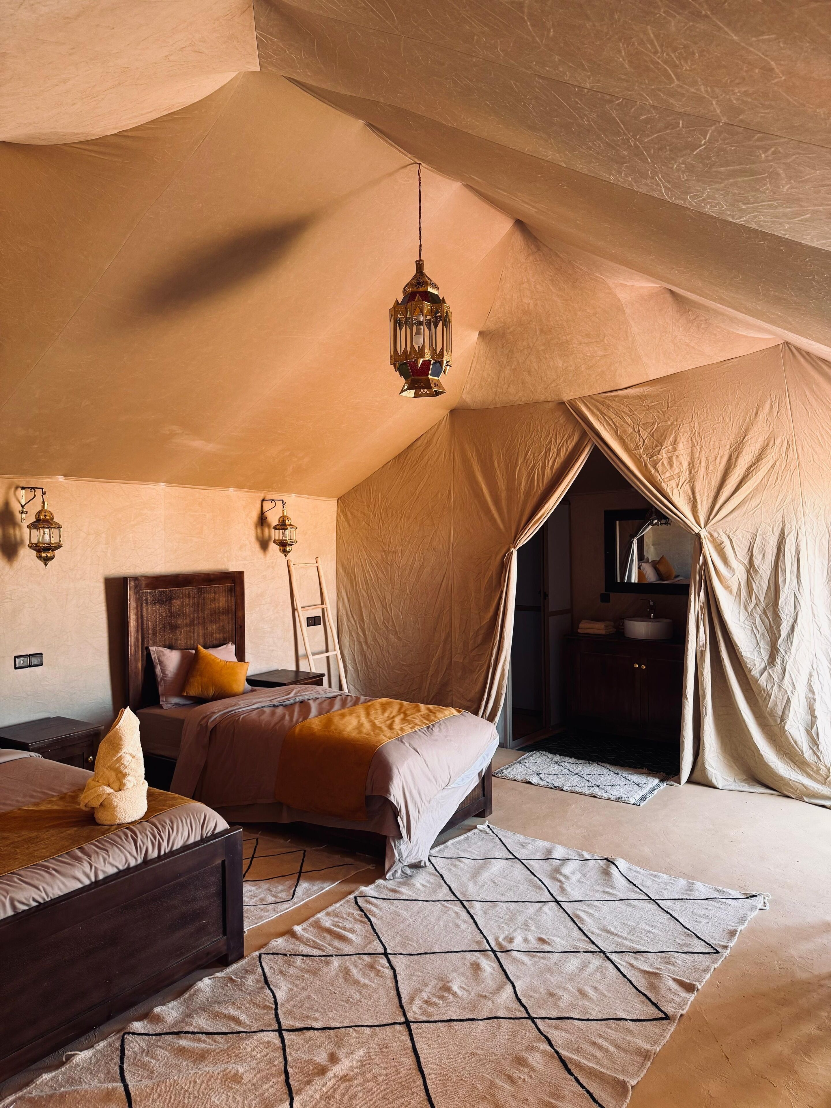 A tented room with two beds, a wooden headboard, a hanging lantern, and a bathroom with a sink and mirror.