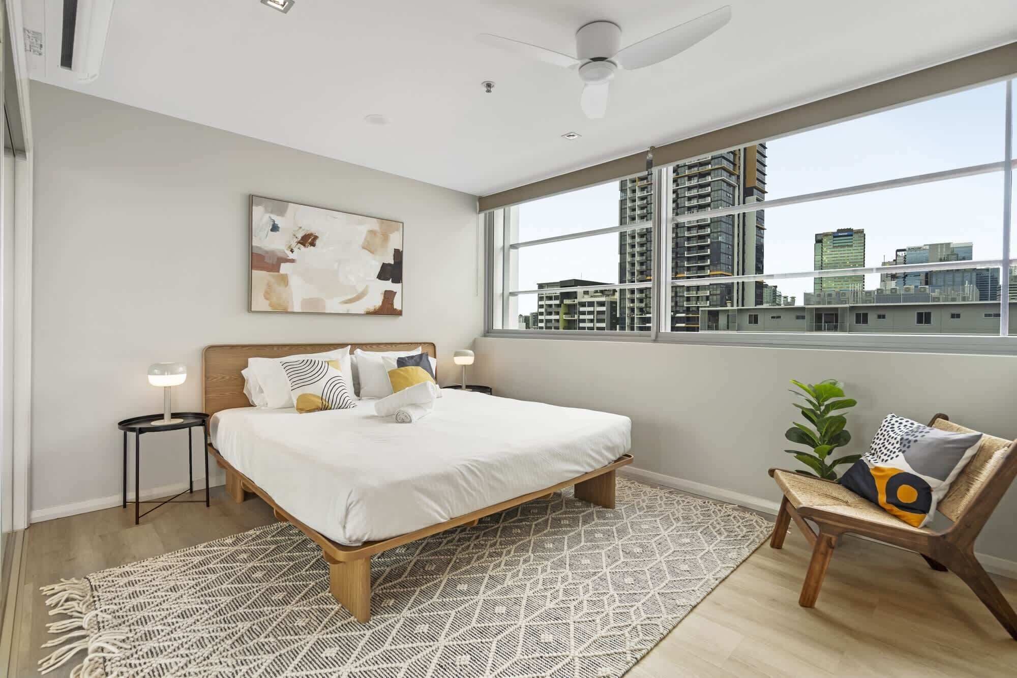 A modern bedroom with a large bed, a city view, a ceiling fan, and a small seating area.