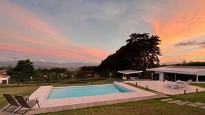 A modern outdoor pool area with a view of the city in the distance, surrounded by greenery and a clear sky. - Room-ao Buena Vista Home - Panoramic Vie (Concepción)