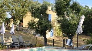 Una piscina con sillas de descanso y sombrillas, rodeada de árboles y un edificio al fondo.