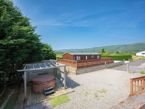 En træhytte med jacuzzi, omgivet af en træterrasse og grusområde, med grønne bakker og en klar himmel i baggrunden.