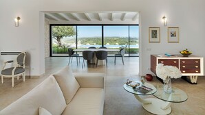 Una sala moderna con un sofá, una mesa de centro de vidrio y un comedor con sillas y una mesa. Hay dos lámparas fijadas a la pared y un mueble con cajones.