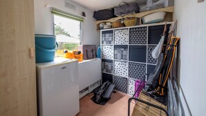 Une cuisine de taille réduite, équipée d’un réfrigérateur, d’un évier et d’une fenêtre. On y trouve des étagères remplies de divers ustensiles de cuisine et un porte-ustensiles contenant des produits de nettoyage.