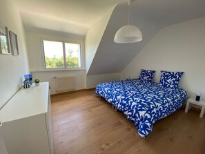 Ein Schlafzimmer mit schräg versiegelter Decke, einem Bett mit blau-weiß gestreifter Bettdecke, einem weißen Nachttisch und einem Fenster mit Blick auf Grünflächen.
