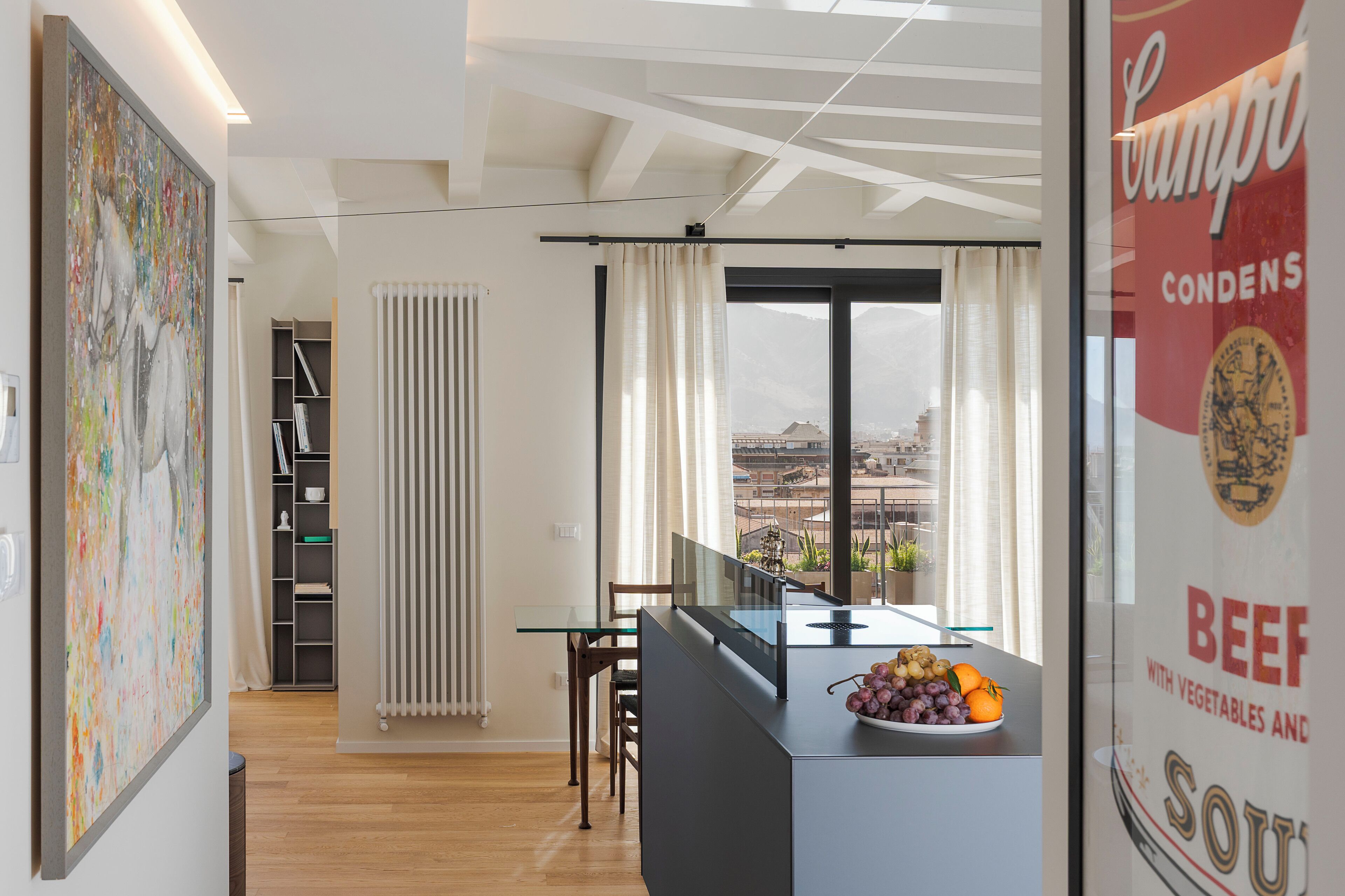 A modern kitchen with a glass island, a dining area with a table and chairs, a radiator, and a view of the city through large windows.