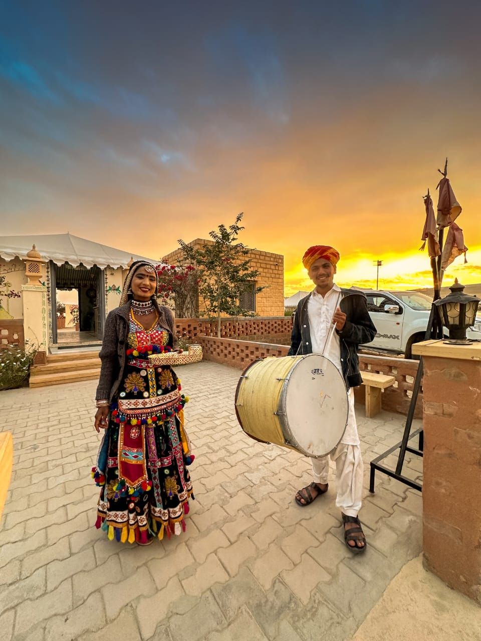 Escena tradicional con una persona tocando un tambor y otra vestida con atuendo colorido, con el atardecer de fondo.