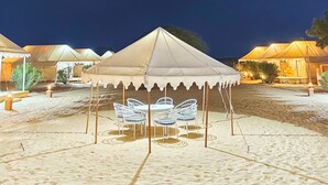 Acampamento no deserto, com tendas, uma área de refeições central com cadeiras de metal branco e chão arenoso.