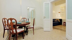 Ein Essbereich mit einem runden Tisch, Holzstühlen und einem gemusterten Teppich. Ein Durchgang führt in ein Wohnzimmer mit einer dunklen Couch und einem kleinen Tisch.