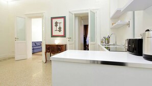Eine moderne Küche mit weißer Arbeitsplatte, einer schwarzen Kaffeemaschine und einem Spülbecken. Ein blaues Sofa und ein gerahmtes Bild sind an der Wand zu sehen.