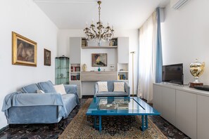 Una sala de estar con un sofá azul, una mesa de centro de vidrio, una araña y un televisor sobre un mueble.